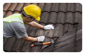 Reparación de Tejados y Cubiertas 1 Tejados Alcala Henares Trabajador con casco revisando las tejas de un tejado con un martillo en mano, destacando la importancia de la reparacion de tejados y cubiertas en edificios